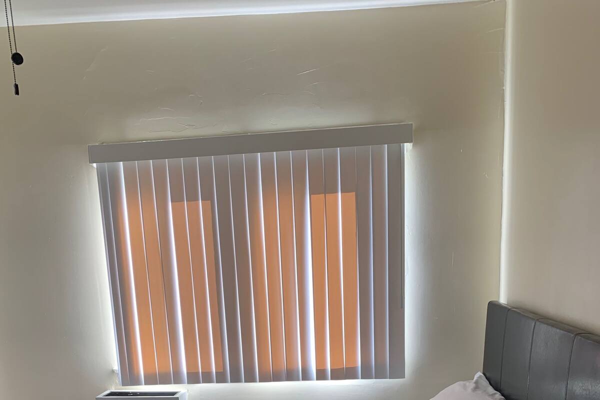 Ein Schlafzimmer mit Bett, weißen Kissen, einem grauen Kopfteil, einem Fenster mit Jalousien und einem kleinen Nachttisch.