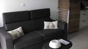 A modern living room with a dark grey sofa, two patterned cushions, a small white table with a bowl and remote, and a framed picture of boats on the wall.