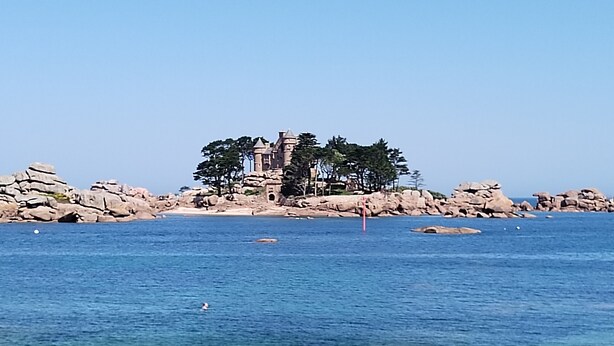 A coastal scene with clear blue water, a sandy beach, and a rocky outcrop with a castle-like structure.