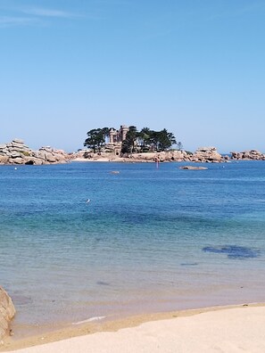 Eine Küstenlandschaft mit klarem, blauem Wasser, einem Sandstrand und einem felsigen Landvorsprung mit einer festungsähnlichen Struktur.