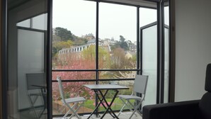 A balcony with a table and chairs, overlooking a garden with pink flowers and a building in the background.