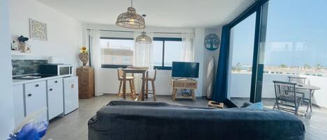 A modern living room with a sofa, a dining area with a table and chairs, a kitchen area with a microwave and refrigerator, and large windows offering a view of the sea.