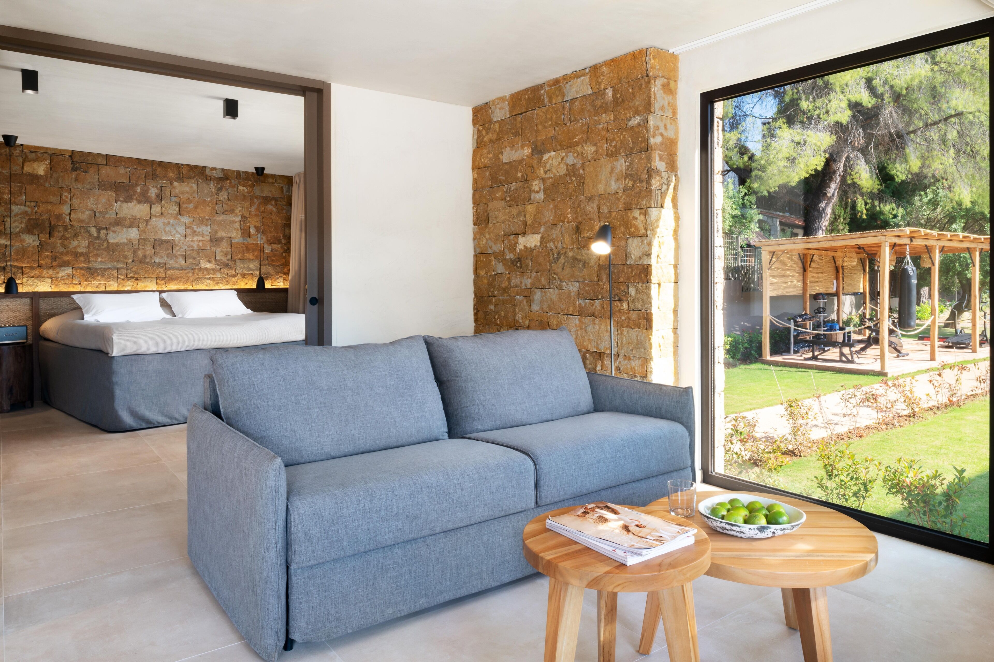 Sala de estar moderna com sofá cinza, mesa de centro de madeira e uma janela grande com vista para um jardim e área de estar externa.