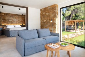 A modern living room with a grey sofa, a wooden coffee table, and a large window with a view of a garden and outdoor seating area. - Amantes Villas and Suites (Sithonia)