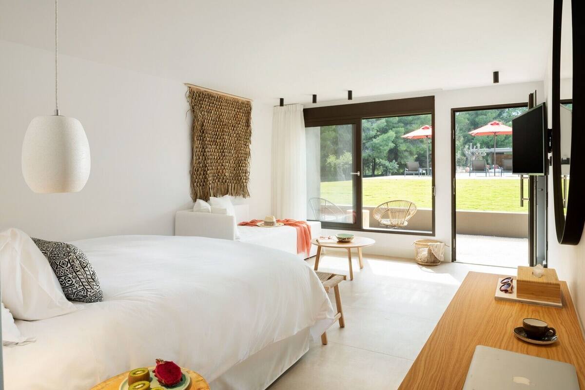 Habitación de hotel moderna con una cama grande, un escritorio con una computadora portátil, un televisor y vistas a un jardín por una puerta corredera.