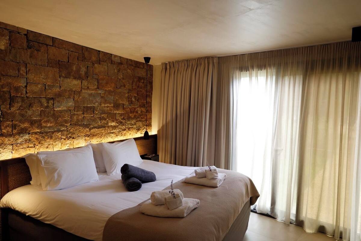 Habitación de hotel con pared de piedra, cama con ropa blanca, cabecero marrón y un ventanal con cortinas.