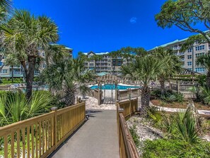 A pathway leading to a pool surrounded by palm trees and a multi-story building with balconies.