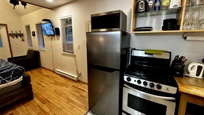 Una cucina compatta con frigorifero, fornello e forno. Si nota uno scaffale con stoviglie e una televisione montata a parete.