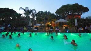 A swimming pool area with people in the water, surrounded by palm trees and umbrellas.