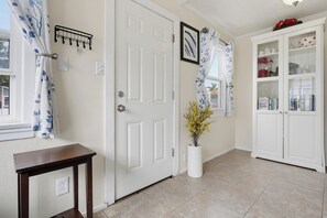 Un pasillo con una puerta blanca, una pequeña mesa, un mueble blanco con puertas de cristal y un jarrón con flores amarillas.