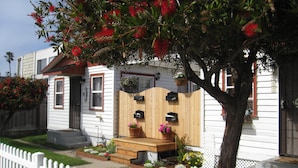 A white house with a wooden front porch, a mailbox, and a small garden with various plants and flowers. - Charming Cottage: 5-minute walk to beach! (San Diego)