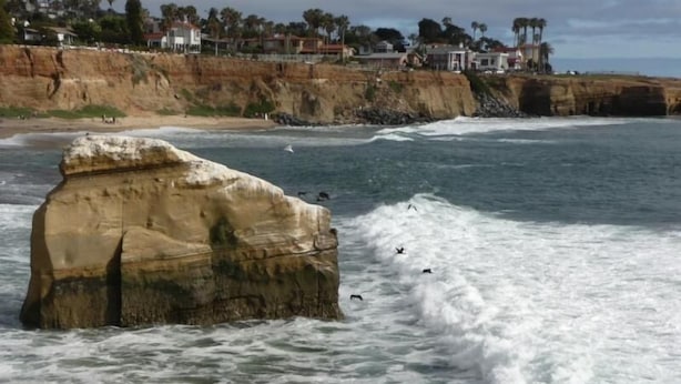 A coastal scene with a large rock formation and waves crashing against it. - Charming Cottage: 5-minute walk to beach! (San Diego)