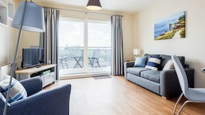 Ein modernes Wohnzimmer mit einem grauen Sofa, einem Fernseher auf einem Holzboden, einer Lampe und einem Gemälde mit einer Küstenlandschaft.