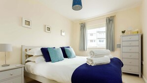 Ein ordentlich bezogenes Bett mit blauer Decke, ein Nachttisch mit Lampe, eine Kommode und ein Fenster mit Vorhängen.