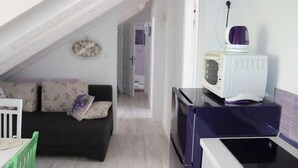 En kompakt opholdsstue med tekøkken, en mørk sofa med mønstrede hynder, et hvidt ur på væggen og et lille spisebord med stole.