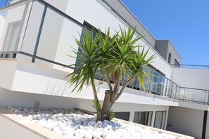 A modern building with a white facade, a balcony with glass railings, and a small tree with green leaves.