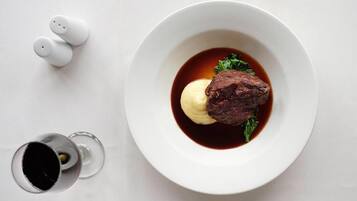 Un piatto bianco con una bistecca e salsa, un bicchiere di vino e dei pepper shaker su una superficie bianca.