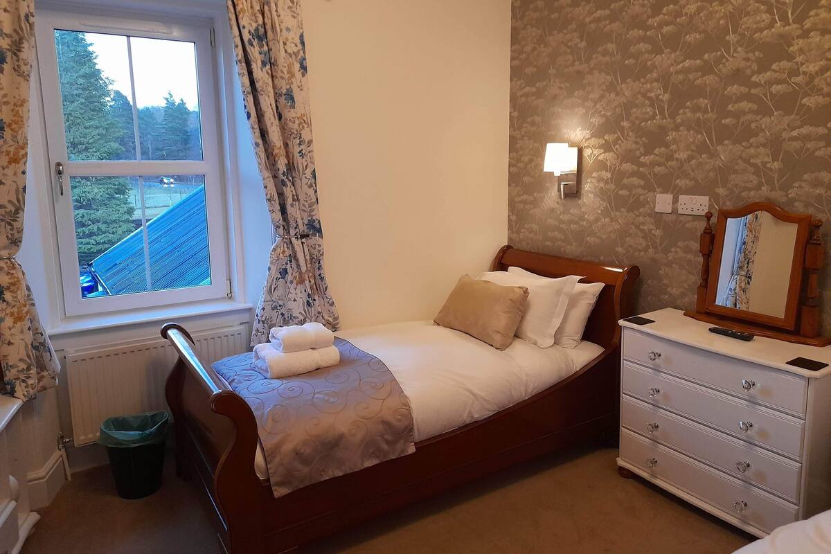 A bedroom with a bed, a dresser, a window with curtains, a radiator, and a wall-mounted light.