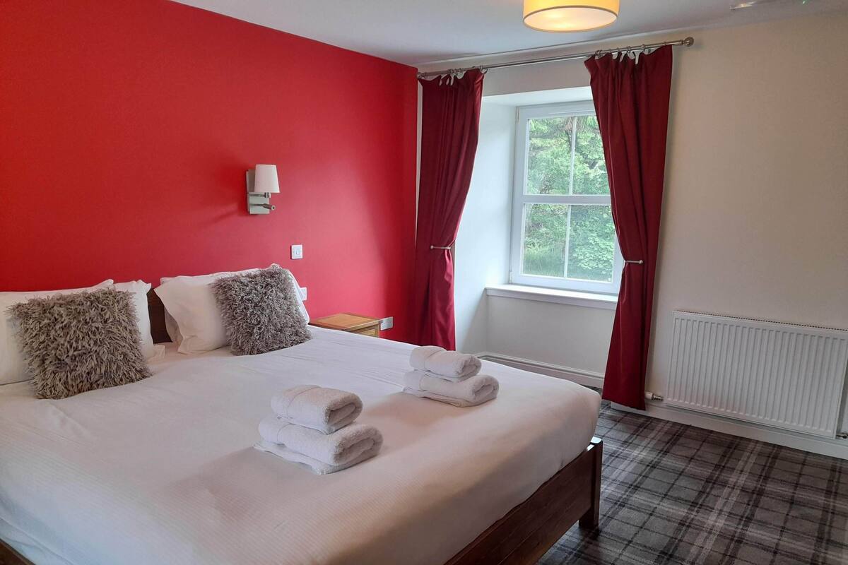 A hotel room with a bed, red walls, a window with curtains, a bedside table, and a lamp.