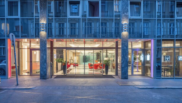 Ein Hotel mit gläserner Fassade, roter Sitzgelegenheit im Inneren und einer Marmorsäule.