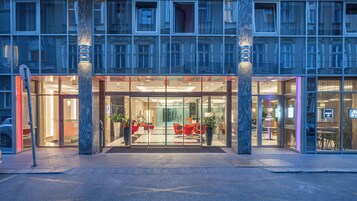 Η είσοδος ενός ξενοδοχείου με μια γυάλινη πρόσοψη, κόκκινα καθίσματα στο εσωτερικό και μια μαρμάρινη κολόνα.