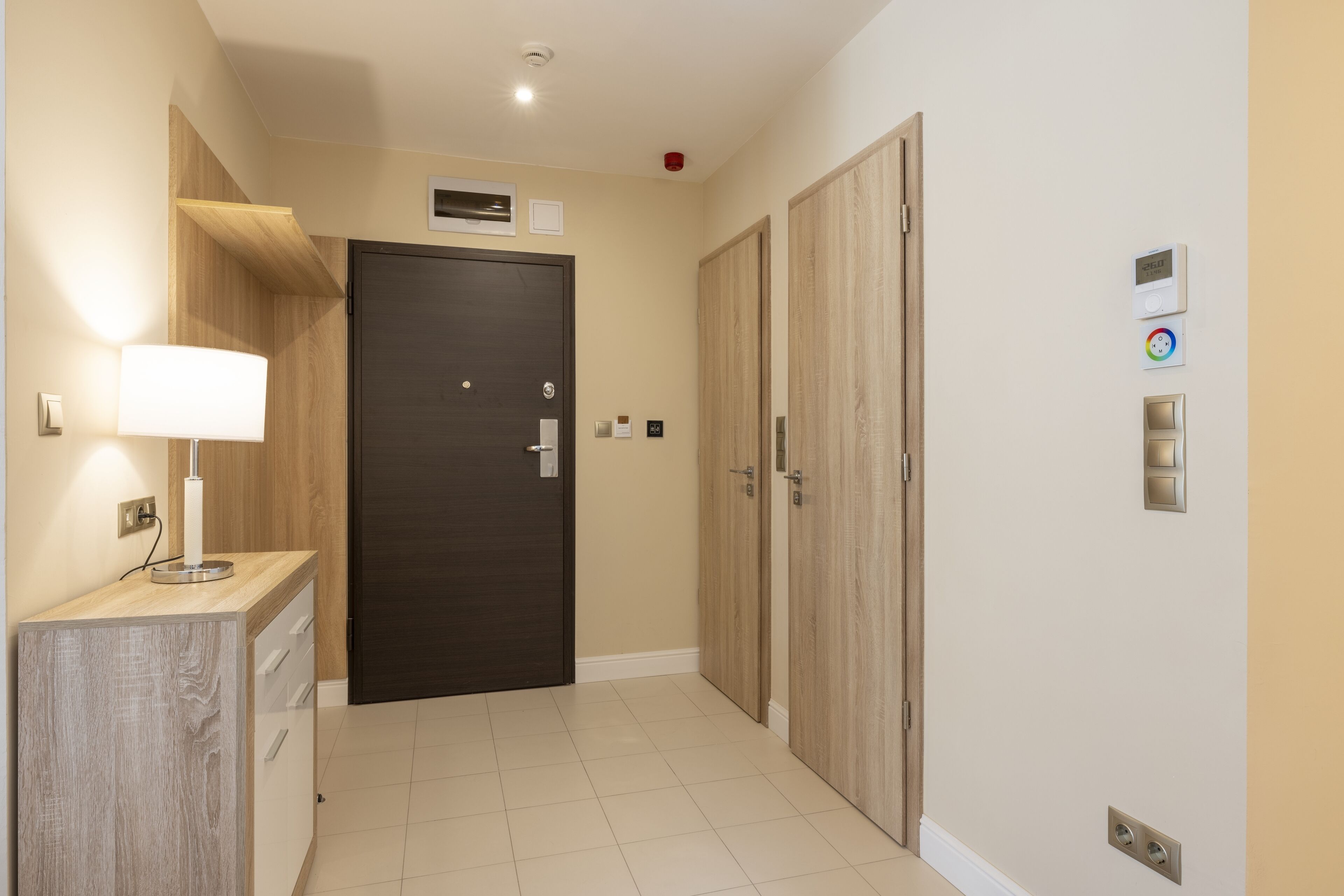 A hallway with a dark wooden door, a small wooden shelf with a lamp, and a wall with a thermostat and a light switch.