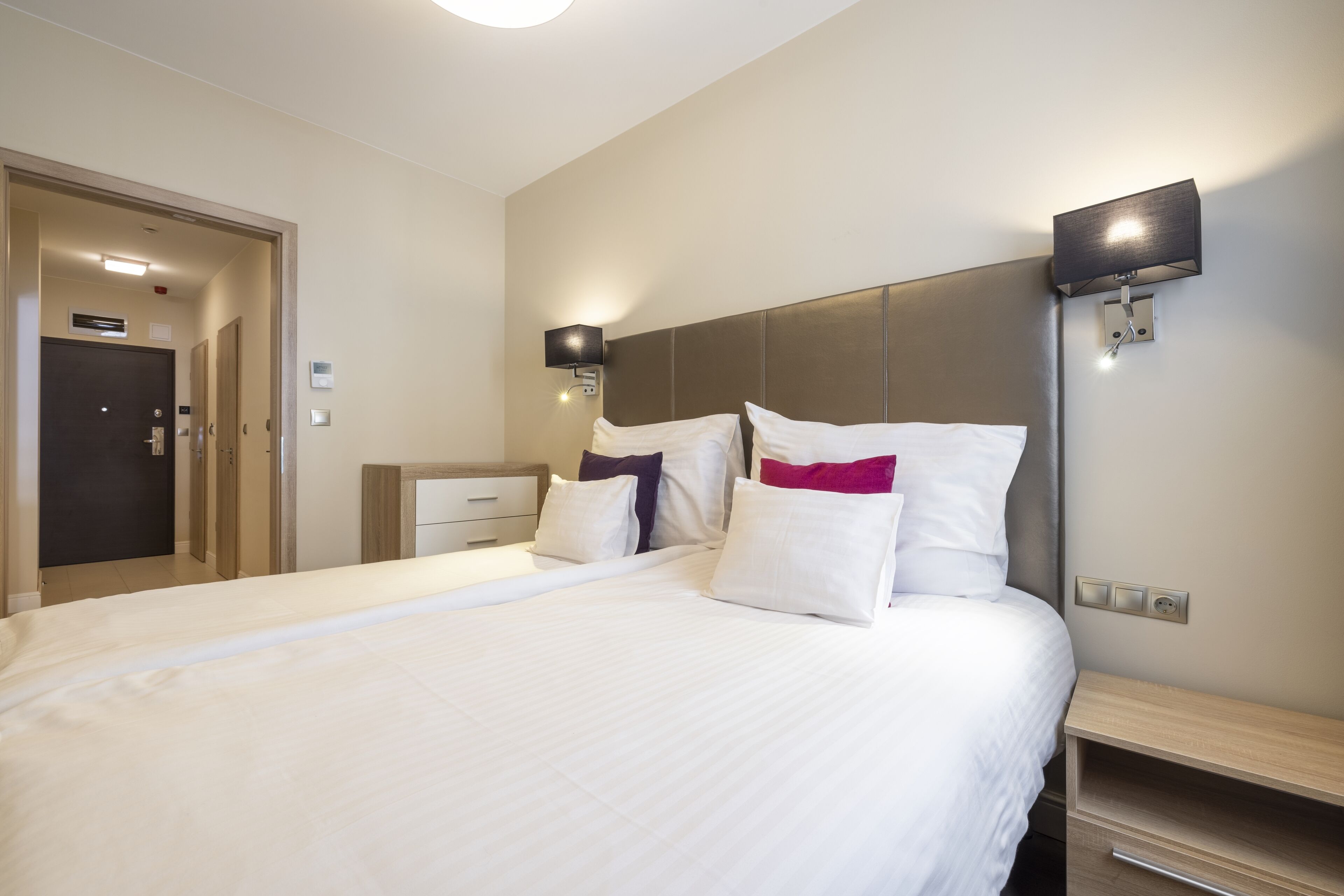 A hotel room with a large bed, bedside tables, a wardrobe, and a wall-mounted light fixture.
