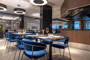 A modern restaurant with round tables and blue velvet chairs, featuring a central bar area and a view of the kitchen. - Silver Crown Hotel & Residence (Budapest)
