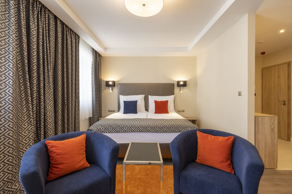 A modern hotel room with a large bed, two bedside lamps, a small table, and two armchairs.