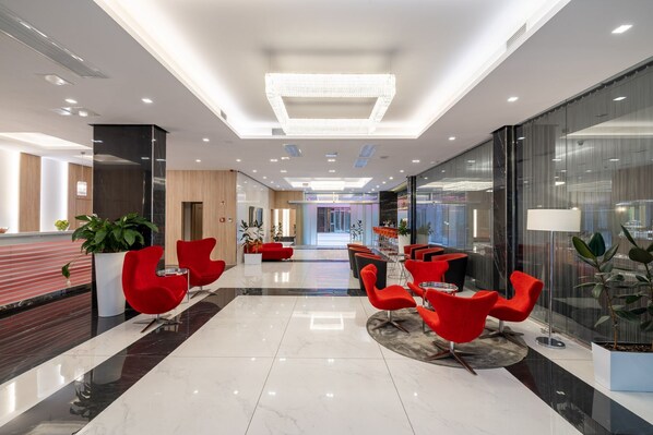 A modern hotel lobby with a marble floor, red chairs, a reception desk, and a large chandelier. - Silver Crown Hotel & Residence (Budapest)