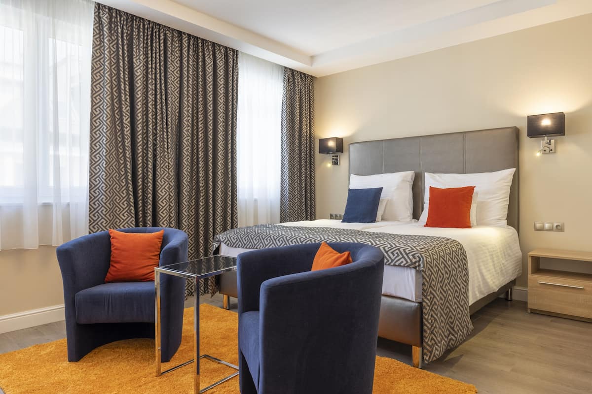 A modern hotel room with a large bed, two armchairs, a glass coffee table, patterned curtains, and wall-mounted lights.