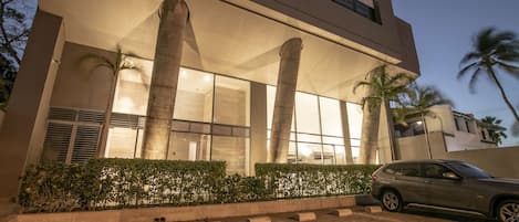Un edificio moderno con grandes columnas, un coche estacionado al frente y palmeras al fondo.