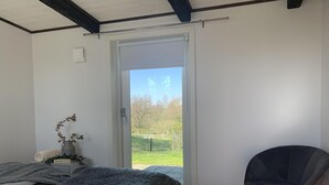 A bedroom with a bed, a chair, a small table with a vase, and a view of a grassy area through a door. - Österlen, fantastic cozy guesthouse for Villa Veranda (Ystad M)