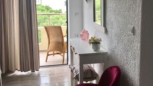 Habitació amb una porta corredissa de vidre que dona a un balcó, un televisor penjat a la paret, una taula petita amb un vas rosa i una planta, una cadira vermella i un aparell d'aire acondicionat blanc.