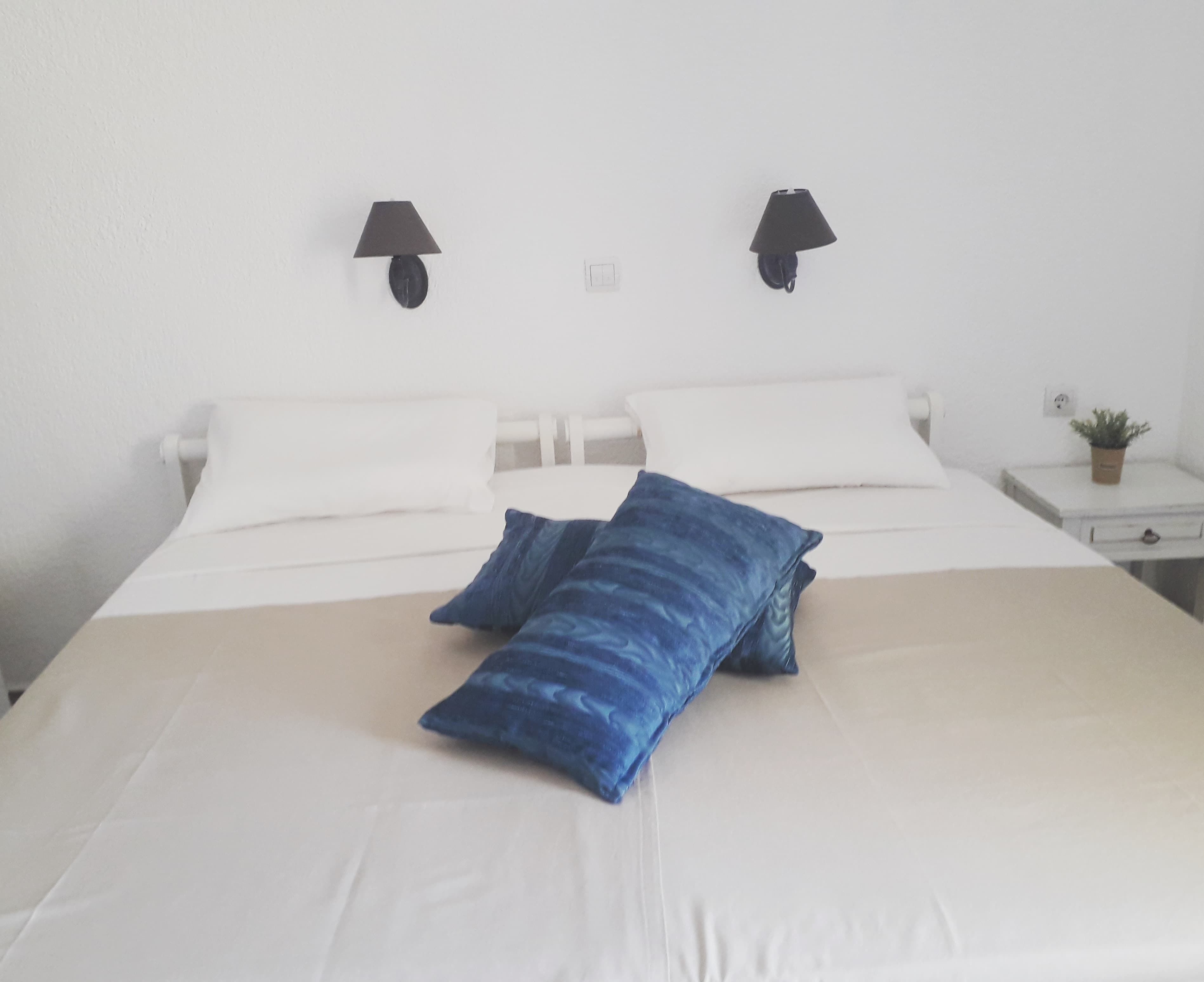 Ein ordentlich bezogenes Bett mit blauem Kopfkissen und weißer Bettdecke. An beiden Seiten des Bettes befinden sich zwei Wandlampen.