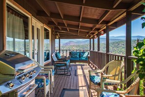 A covered outdoor deck with a grill, wicker chairs, and a view of mountains.