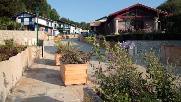 A courtyard with stone pathways, wooden planters, and a building with a red roof.