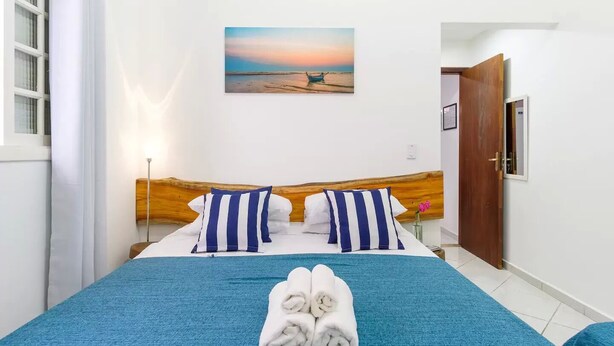 A neatly made bed with blue bedding and striped pillows, a pair of towels, and a painting of a beach scene on the wall. - At Casa em Ubatuba - Praia do Itaguá, excellent for families and long stays. (Ubatuba)