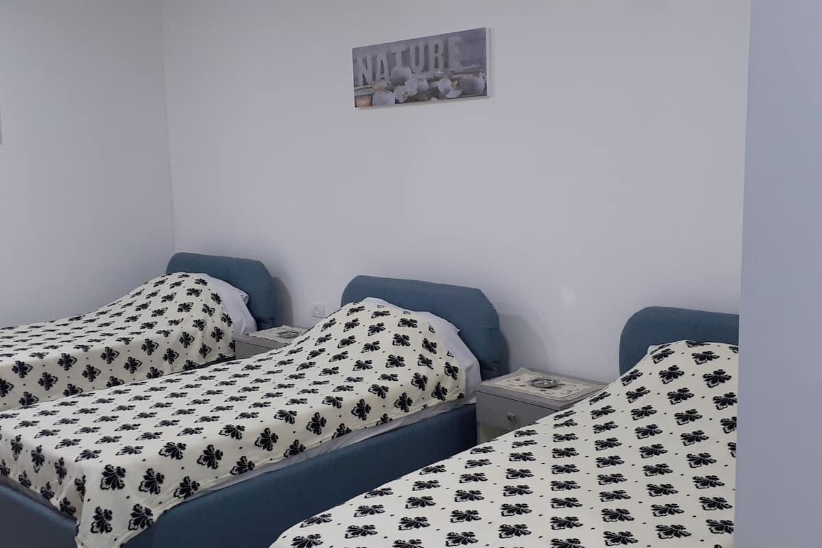 A room with three single beds, each with a blue headboard and a floral-patterned bedspread. There are two small bedside tables with lamps, and a wall-mounted picture frame above the beds.