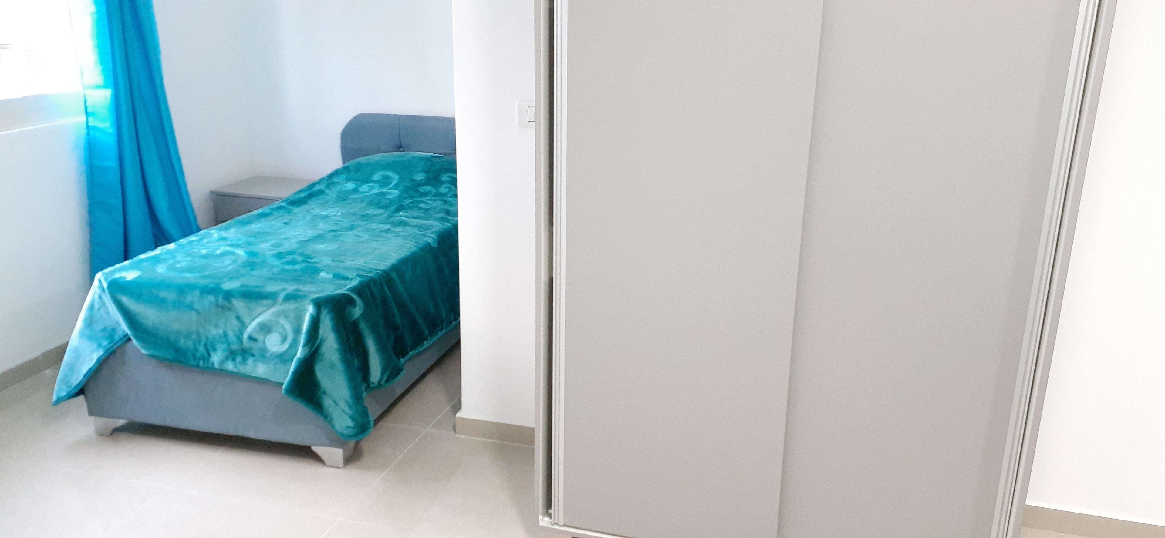 A bedroom with a bed covered in a teal bedspread, a grey sofa, and a small bedside table.