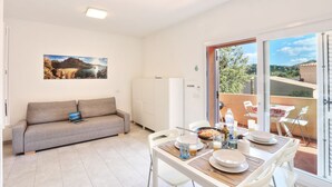 Ein modernes Wohnzimmer mit einer Couch, einem Esstisch und Stühlen. Ein Kühlschrank, ein Gemälde an der Wand und ein Blick ins Freie durch große Fenster sind ebenfalls zu sehen.