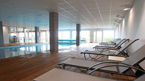Zona de piscina interior con suelo de madera, sillones de descanso y vistas a la piscina.