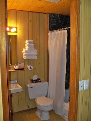 A wooden bathroom with a toilet, sink, and stacked towels. - Stardust Motel (Haines Junction)