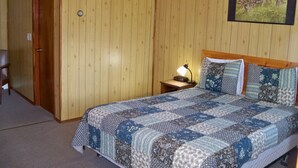 A bedroom with a wooden paneled wall, a bed with a patterned quilt, a nightstand with a lamp, and a framed picture of wolves.