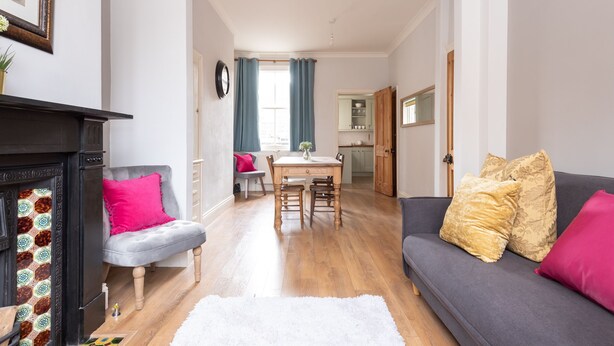 En stue med pejs, en grå sofa med pink puder, et spisebord med stole og et køkken i baggrunden.