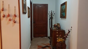 Um corredor com uma porta de madeira, um quadro emoldurado, um vaso com flores e um tapete estampado.