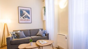 A living room with a grey sofa, two small round tables, a framed poster on the wall, a floor lamp, and a radiator. - Le Saint Nicolas - One Bedroom Apartment, Sleeps 4 (Toulouse)