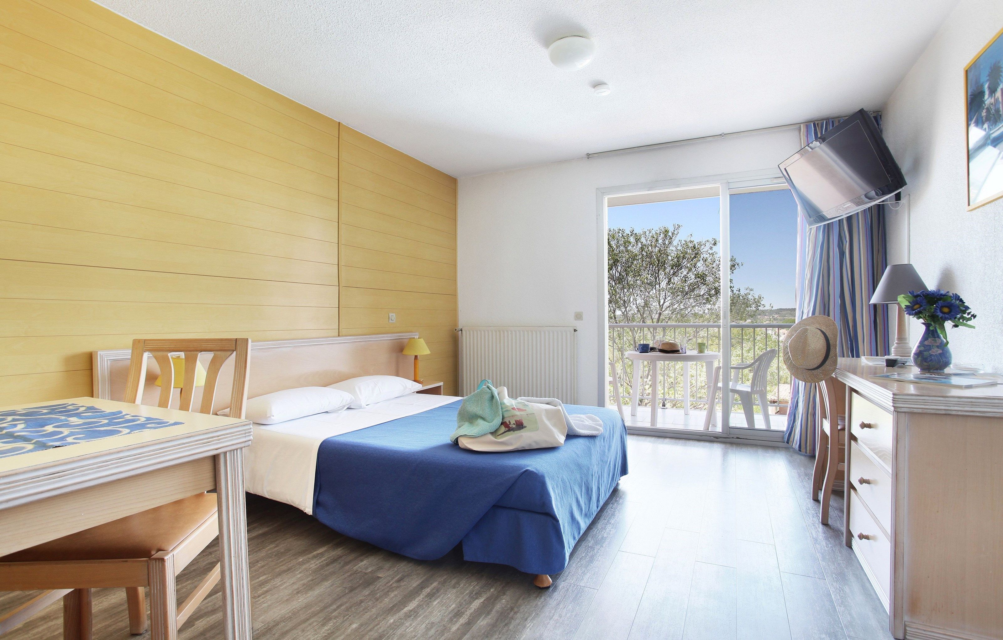 A hotel room with a bed, a desk, a chair, a lamp, a dresser, and a balcony with a view.