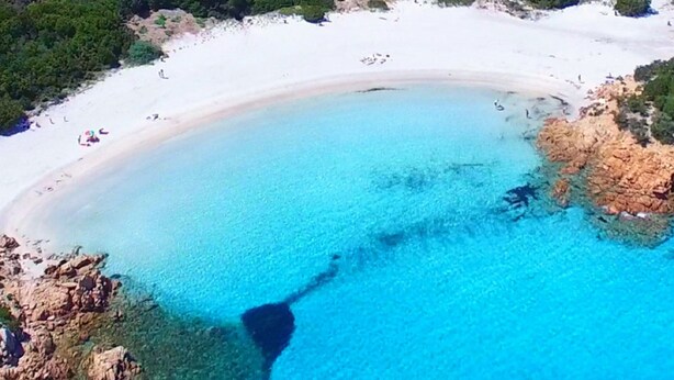 Vista aérea de una playa con aguas turquesas cristalinas, arena blanca y afloramientos rocosos.