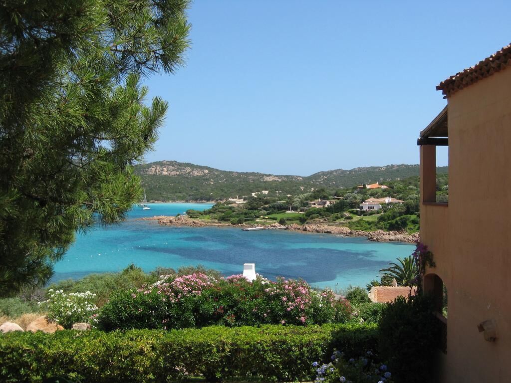 A coastal view with a clear blue sea, green hills, and a building with a tiled roof.
